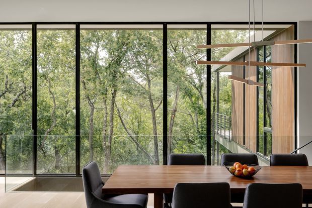 A contemporary dining area with a long timber dining table and dark upholstered chairs, positioned beside expansive floor-to-ceiling black-framed glazing. The glazing overlooks dense leafy vegetation and reflects the surrounding interior. A sculptural linear pendant light with timber-toned horizontal elements hangs above the table, while vertical timber wall panelling and an exterior balcony are visible to the right. The room has a calm, nature-connected atmosphere and a refined contemporary aesthetic.