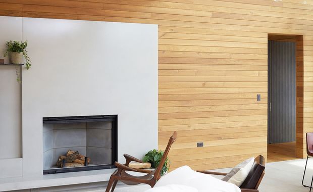 A warm, refined living room featuring a timber-lined ceiling and wall, a minimalist white fireplace surround with an inset firebox, and mid-century-style wooden lounge chairs around a low seating area. A dark ribbed doorway opens into an adjacent space, with simple plants and neutral furnishings adding a calm, natural feel.