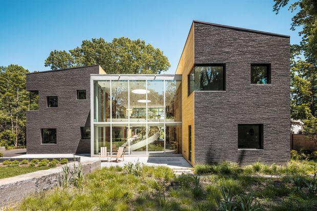 Exterior view of a contemporary two-storey residence composed of two dark brick-clad volumes connected by a transparent central glazed atrium. The central glazing reveals a bright interior with a staircase and circular pendant lights. A landscaped garden and paved path lead toward the entrance, with mature trees surrounding the property.