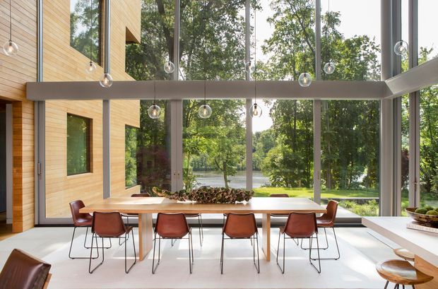 A bright, contemporary dining area with a long timber dining table and modern leather dining chairs, set within a double-height glazed space. Full-height windows provide views to a leafy wooded landscape and water beyond. Warm timber wall cladding, slim grey structural frames, polished light flooring, and suspended globe pendant lights create a refined indoor-outdoor connection.