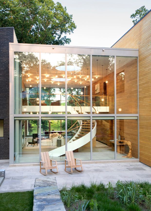 Exterior view of a contemporary multi-level home centered on a tall glazed courtyard facade. The floor-to-ceiling window wall reveals a bright interior with a sculptural curved staircase, timber wall cladding, extensive pendant lighting, and open-plan seating areas. Pale stone paving, two timber rocking chairs, landscaped planting, and a low stone garden wall occupy the foreground. Dark brick and warm timber-clad exterior volumes frame the glazed central section.