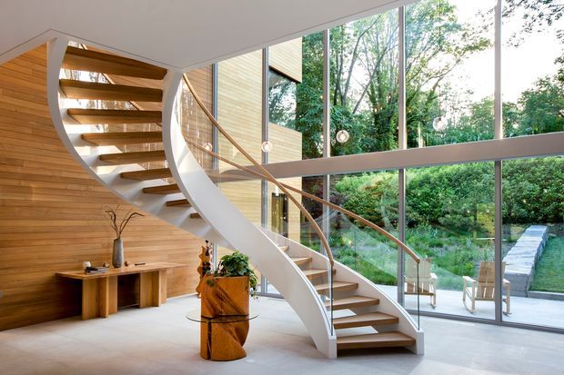 A bright, contemporary double-height living space dominated by a sculptural curved staircase with open timber treads, white stringers, and a slender timber handrail. Warm horizontal timber wall panelling lines the left side, while extensive floor-to-ceiling glazing overlooks a leafy garden and paved path. Minimal furnishings, pale flooring, decorative timber elements, and pendant lights create an airy, refined interior with strong indoor-outdoor connection.