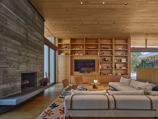 Warm, mid-century-inspired living room with a large built-in timber entertainment and shelving wall, a linear fireplace set into a dark textured feature wall, full-height glazing, timber-lined ceiling, and a neutral sectional sofa arranged around a low coffee table.