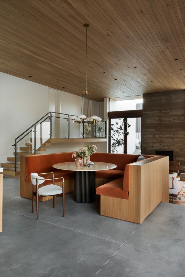 A refined open-plan dining and living area with a built-in rust-orange upholstered banquette surrounding a round dining table. The room features a warm timber-lined ceiling, polished concrete floor, dark timber feature wall, timber stair and balustrade, expansive glazing, and a sculptural suspended chandelier. The space has a contemporary, warm modern character with strong natural material contrast.