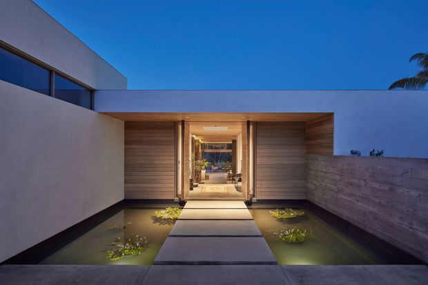 Twilight exterior view of a minimalist modern residence entrance. A broad concrete stepping-stone path crosses shallow reflecting pools with aquatic planting and leads through a warm timber-lined portal into a glazed interior. Smooth rendered walls, horizontal timber cladding, concealed lighting, and a flat roof create a calm, geometric composition.