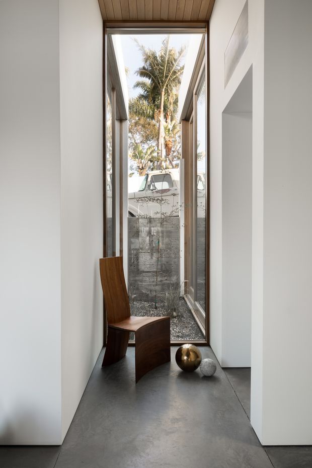 A narrow, minimalist interior passage with a full-height glazed opening at the end, looking onto a small planted courtyard and palm trees. The passage has smooth white walls, a dark polished concrete floor, a warm timber slatted ceiling, slender timber-framed glazing, and a sculptural curved timber chair with metallic decorative spheres.
