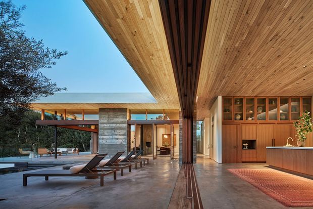 A sophisticated open-plan indoor-outdoor living space beside a pool, framed by expansive glazing and dramatic cantilevered roof planes. Warm timber lines the soffit and ceiling, with matching built-in cabinetry and a kitchen island. Polished concrete flooring, dark slender framing, integrated lighting, and a long view toward the landscaped exterior create a refined contemporary architectural composition.