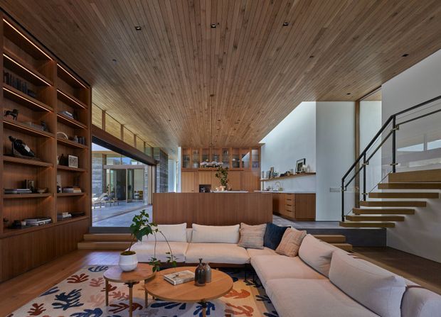 A spacious contemporary living room with an expansive warm timber-lined ceiling, integrated dark timber shelving, a large modular sectional sofa, and a pair of round timber coffee tables. The open-plan space includes a timber kitchen or bar joinery zone in the background and a floating staircase with black metal balustrade at right. Glazed openings connect the room to an outdoor area, while recessed ceiling lighting and built-in cabinetry create a refined, cohesive interior.