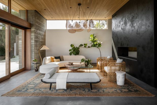 A refined contemporary living room with expansive floor-to-ceiling glazing, a warm timber-lined ceiling, polished concrete flooring, and a dramatic dark textured feature wall incorporating a fireplace and television. The room is furnished with a large light-coloured sectional sofa, woven rattan armchairs, a low coffee table, patterned area rug, indoor plants, and sculptural pendant lighting. The space feels airy, warm, and sophisticated, with strong indoor-outdoor connection.