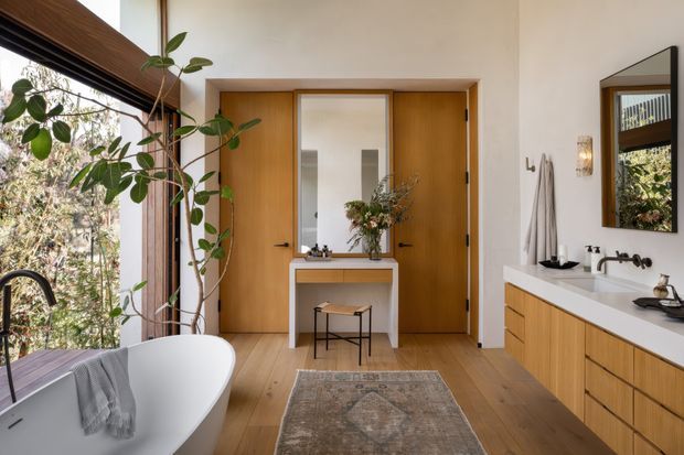 Bright contemporary bathroom with a freestanding white bathtub beside a large window, a long timber vanity with a white countertop and wall-mounted faucets, and a timber-framed mirrored cabinet. Warm timber flooring, timber doors, indoor plants, and a small console with stool create a refined, natural interior. The mirror reflects additional glazing and greenery.