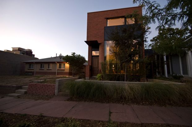Twilight street-level exterior view of a contemporary multi-storey urban residence. The home has a strong rectilinear form, exposed reddish-brown brick facade, dark metal or painted framing, recessed openings, and a tall glazed entry or living area partially screened by landscaping. A low brick retaining wall, concrete footpath, and neighbouring houses are visible in the foreground and background.