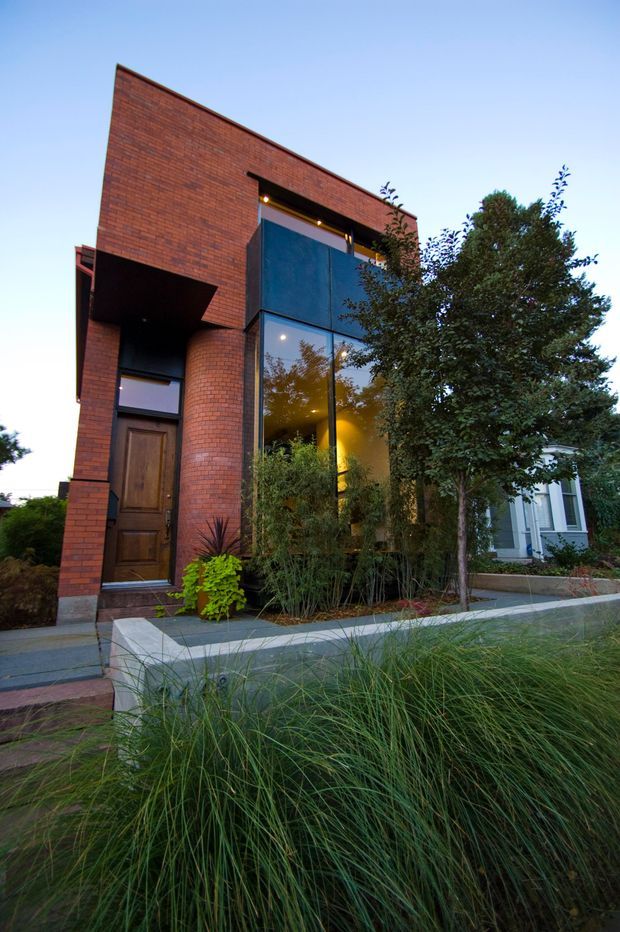 Street-level exterior view of a narrow contemporary brick residence. The home features a tall, strongly geometric red-brick facade, a projecting dark recessed entry canopy, timber front door, and large floor-to-ceiling glazed openings revealing warm interior lighting. A raised concrete planter and layered grasses, shrubs, and trees form the landscaped front edge, with a neighbouring light-coloured building visible to the side.