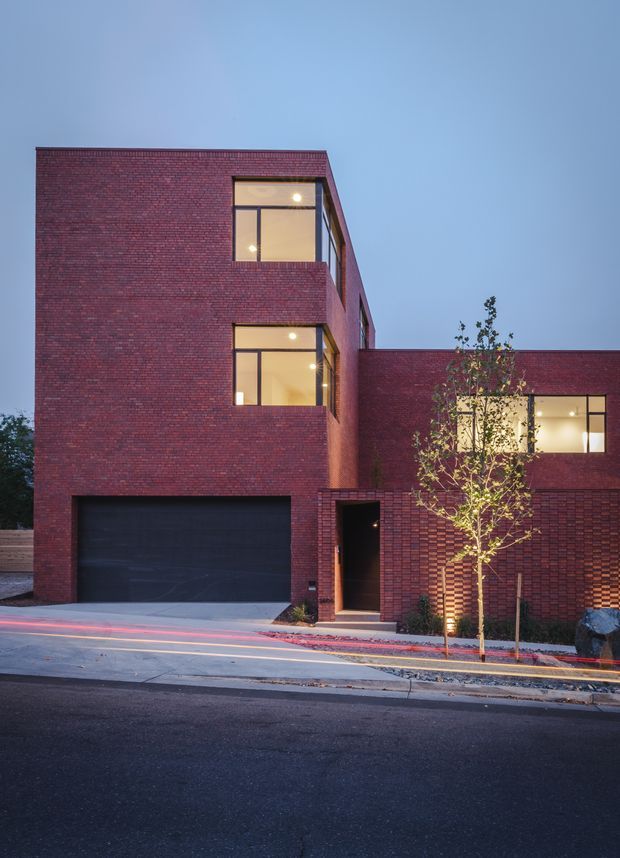 Exterior street-level view of a contemporary multi-storey brick residence at dusk. The building has a strongly rectilinear massing, deep red brick walls, large dark-framed windows with warm interior lighting, an integrated dark garage door, and a recessed entry. A lower wing extends to the right, with a young street tree and paved footpath in the foreground.