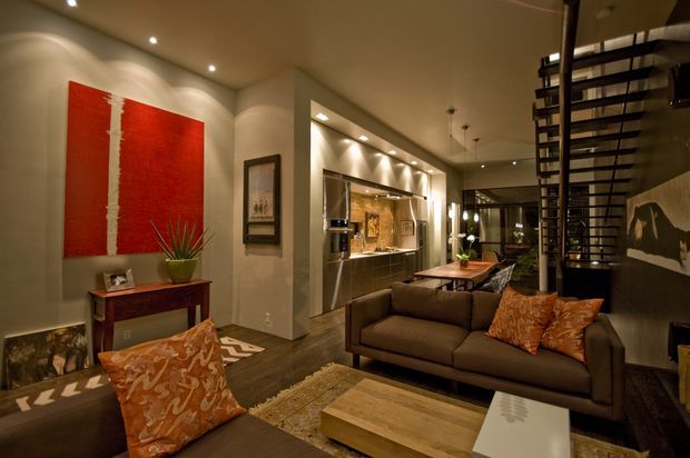 Warmly lit contemporary open-plan living room with a dark upholstered sofa, patterned orange cushions, timber coffee table, console table, area rug, and large red abstract artwork. The room opens toward a dining area and a sleek kitchen with integrated cabinetry and a reflective splashback. A dramatic black open-riser stair or screened stair structure forms a strong architectural feature on the right, with recessed ceiling lighting throughout.