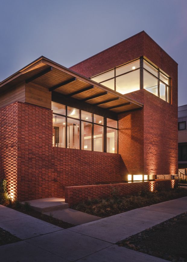 Twilight exterior view of a contemporary multi-level brick house. The building features deep red brick walls, large dark-framed glazed openings, warm interior lighting, and projecting timber-lined roof and canopy elements. Concrete footpaths and low landscaped planting surround the street-facing facade.