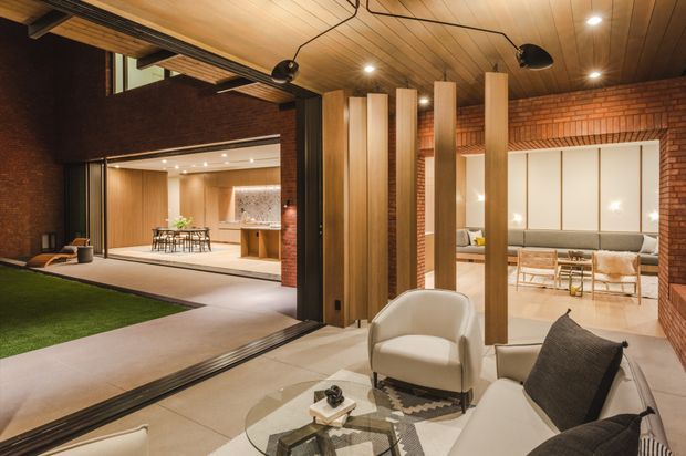 A sophisticated open-plan residential interior centered on a covered outdoor courtyard or terrace, with large retractable glazed openings connecting it to the living and dining areas. The space features exposed red brick walls, warm timber-lined ceilings, vertical timber screens, polished concrete or large pale tiled flooring, and contemporary built-in lighting. A lounge seating area occupies the foreground, while a dining space and kitchen are visible beyond. The design has strong indoor–outdoor connection and warm architectural detailing.