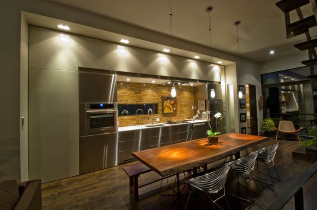 A spacious contemporary kitchen and dining area with a long wall of sleek metallic cabinetry, integrated stainless-steel oven, dark benchtops, and a warm brick-look tiled splashback. A large rustic timber dining table with wire-frame chairs occupies the foreground, while pendant lighting, recessed cabinetry lights, and dark timber flooring create a warm, industrial-modern atmosphere. An open stair structure and adjacent living area are visible at the right.