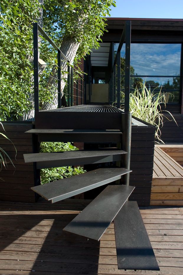 Exterior view of a contemporary dwelling with a dark timber-clad facade, large reflective glazing, and a sculptural open-riser metal staircase leading up from a timber deck. Dense planting surrounds the elevated entry, with dark steel posts and wire balustrades defining the approach.