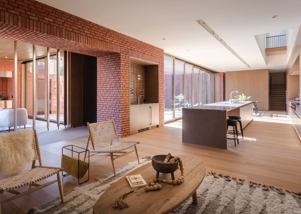 Open-plan contemporary home interior combining a living room and kitchen. The space features exposed red brick walls and arched brick detailing, full-height glazing, light timber flooring, a dark kitchen island, timber slatted joinery, and mid-century-inspired lounge furniture. A stairway and upper-level void are visible in the background.