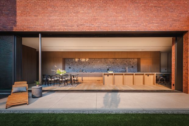 A contemporary indoor-outdoor kitchen and dining space opening onto a paved terrace. The broad dark-framed opening is set within a red brick facade, with a timber-lined ceiling and walls, a long timber kitchen island and cabinetry, dark stone splashback, dining table and chairs, and built-in workspace cabinetry at the right. A small outdoor planter and lounge chair sit on the terrace in the foreground, with lawn beyond.