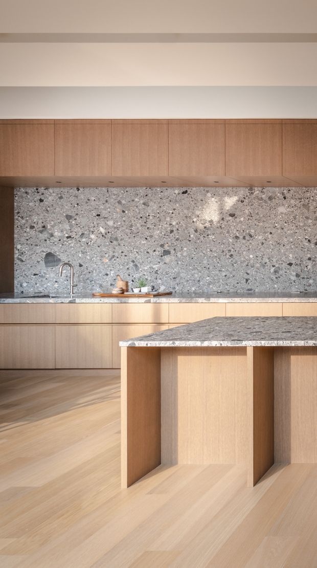 A refined contemporary kitchen featuring extensive light timber cabinetry, a large grey speckled stone island and matching backsplash, integrated storage, a minimalist sink and tap, and wide-plank timber flooring. Warm natural light creates a calm, cohesive atmosphere.
