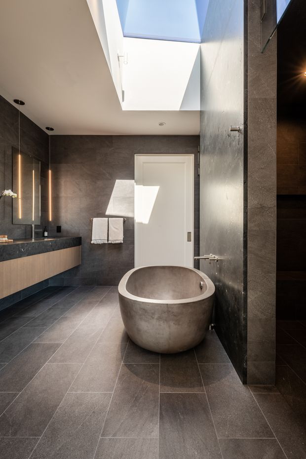 Contemporary bathroom with a freestanding round concrete bathtub, dark stone-look tiled walls and floor, and a floating timber vanity with a dark benchtop. A large skylight brings strong natural light into the space, while a frameless glass shower enclosure is visible at right. The room has clean lines, integrated lighting, and a restrained dark neutral palette.