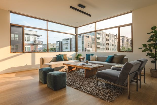 A bright, spacious contemporary living room with full-height corner glazing overlooking an urban residential setting. The room features pale timber flooring, warm off-white walls, a large neutral sectional sofa, charcoal lounge chairs and ottomans, teal cushions, a textured shag rug, and a substantial rustic timber coffee table. Minimal black ceiling-mounted lighting and a tall indoor plant complete the refined, airy interior.