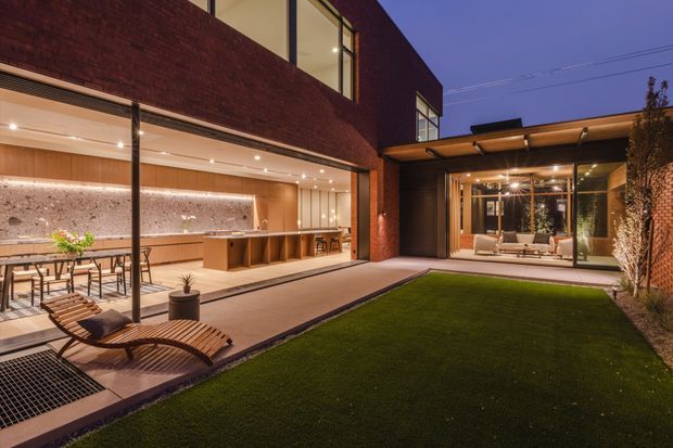 Twilight view of a refined contemporary brick residence arranged around a landscaped courtyard. The open-plan kitchen and dining area is visible on the left beneath a deep overhang, while a glazed living room opens to the courtyard on the right. Warm recessed lighting, timber cabinetry, stone surfaces, extensive glazing, paved terraces and a covered outdoor lounge create an inviting indoor-outdoor connection.