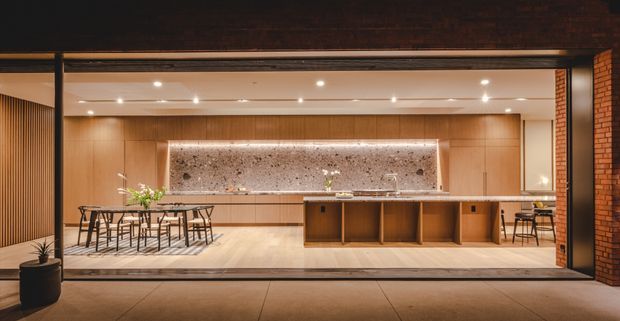 A warmly lit contemporary open-plan kitchen and dining space viewed from an outdoor terrace through fully open, dark-framed glazing. The room features extensive timber-toned cabinetry, a large island with open shelving, a dramatic stone feature splashback with concealed lighting, a dining table with chairs, recessed ceiling lights, and exposed brickwork at the sides. The space has a strong indoor-outdoor connection and a refined, inviting atmosphere.