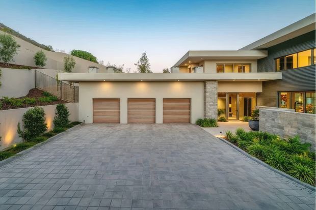 Contemporary multi-level home exterior viewed from a broad paved driveway. The facade combines smooth light render, dark horizontal cladding, stacked-stone sections, warm recessed lighting, large glazed entry areas, and three integrated garage doors. Landscaped garden beds and retaining walls flank the driveway on a sloped suburban site.