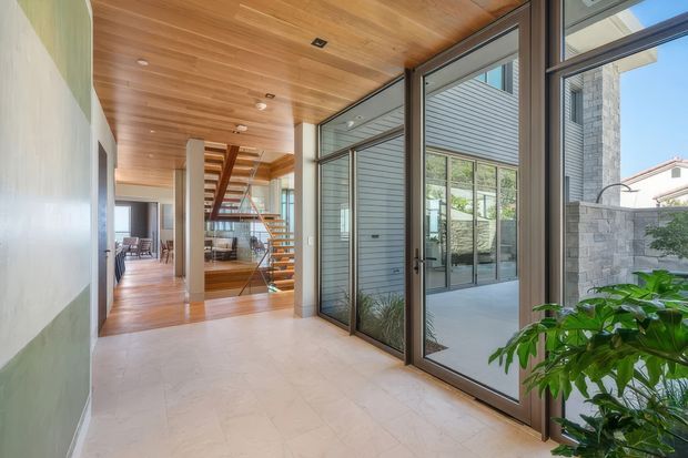 A bright contemporary residential entry hall with large floor-to-ceiling glazed doors opening to an outdoor courtyard. The space features a warm timber-lined ceiling, pale stone or tile flooring, exposed structural columns, dark metal-framed glazing, and a floating timber staircase leading toward an open living area. A leafy indoor plant and courtyard landscaping soften the clean architectural palette.