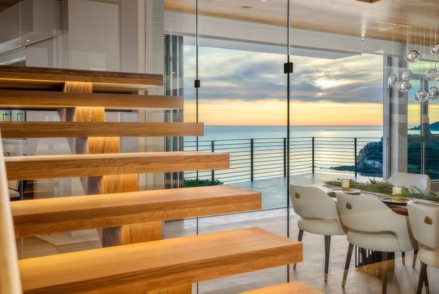 Contemporary open-plan dining and stair area overlooking the ocean at sunset. A sculptural open-riser timber staircase dominates the foreground, while a glass wall and balcony railing provide expansive coastal views. The dining area includes a light-coloured table, upholstered chairs, and a cluster of spherical pendant lights.