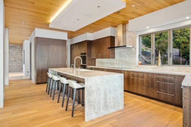 A bright, spacious contemporary kitchen with a large waterfall-edge stone island and seating for four. The kitchen features dark timber cabinetry, a matching timber-lined ceiling, warm timber flooring, a pale textured stone benchtop and splashback, integrated appliances, and large windows overlooking greenery. A hallway and adjoining areas are visible beyond the kitchen.