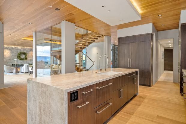 A spacious, open-plan contemporary kitchen centered on a large waterfall-edge island with a pale stone benchtop, dark timber cabinetry, integrated appliances, and a gooseneck tap. Warm timber ceiling and flooring continue through the home, while frameless glass partitions reveal a living area and floating staircase beyond. The room has abundant natural light, refined built-in joinery, and a strong connection between kitchen, circulation, and living spaces.