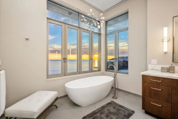 A bright contemporary bathroom with a freestanding white oval bathtub positioned in front of large corner windows overlooking a sunset water view. The room includes a white upholstered bench, wall-mounted vertical sconces, a dark timber vanity with a white stone benchtop, grey flooring, and a modern sculptural pendant light.