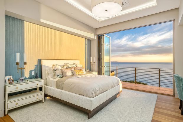 A bright, refined bedroom with a large upholstered bed, pale bedding, bedside lighting, a white drawer console, and a soft area rug. The room opens to a timber-floored balcony or terrace with a metal balustrade and expansive ocean views. A blue and warm beige feature wall, layered ceiling detail, and generous glazing add a contemporary coastal feel.
