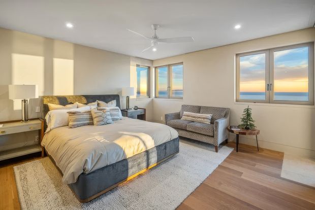 Bright, comfortably furnished bedroom with a large upholstered bed, bedside tables, patterned armchair, rug, timber-look flooring, neutral beige walls, and expansive windows overlooking a coastal horizon. Recessed lighting and a ceiling fan contribute to a relaxed, contemporary feel.