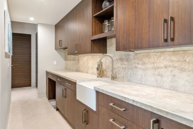 A compact, modern galley kitchen with extensive warm wood cabinetry, pale stone-look benchtops, a white apron-front sink, brushed metal hardware, and a coordinated textured stone-look splashback. Recessed ceiling lighting and a light floor create a bright, clean space, with a hallway visible at the rear.