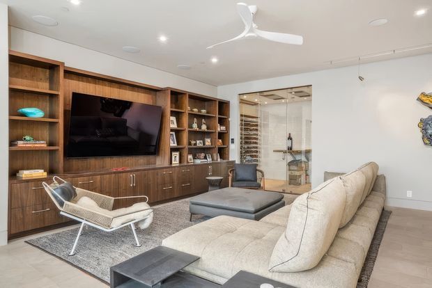 Contemporary living room with extensive built-in dark timber cabinetry and open shelving surrounding a wall-mounted television. A large light sectional sofa and ottoman sit on a textured grey rug, with a sculptural lounge chair in the foreground. Glass doors reveal a compact wine storage room, while recessed lighting and a white ceiling fan create a bright, refined interior.