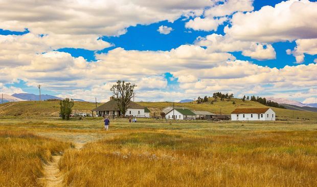 Wide rural landscape showing a small cluster of simple white farm buildings with pitched roofs spread across golden grassland, with rolling hills and mountains in the distance beneath a dramatic partly cloudy blue sky.