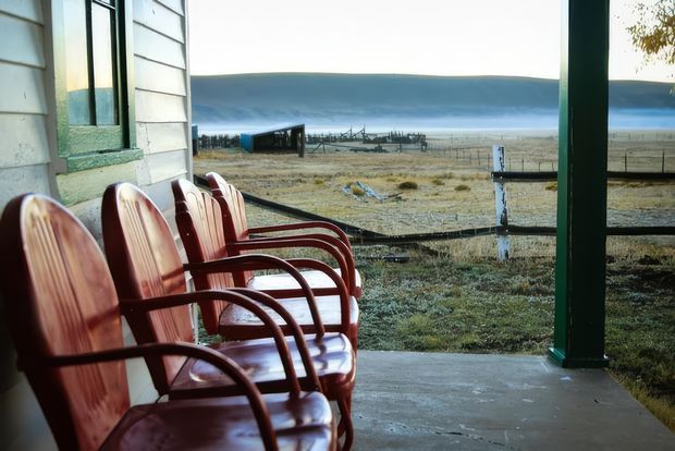 A covered rural verandah beside a weatherboard building, furnished with a row of red wooden armchairs. The view looks across fenced open farmland toward a broad body of water and distant shoreline, creating a quiet, remote setting.