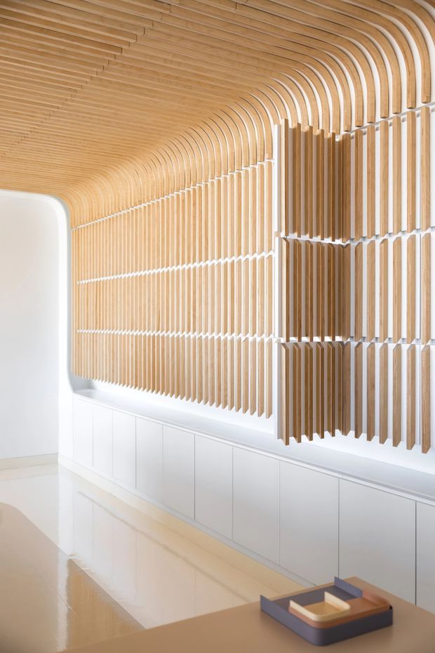 A bright, refined interior corridor or reception-like space featuring an extensive sculptural wall and ceiling installation of curved and vertical light timber slats. The slats create a continuous ribbed, arched form, with white integrated cabinetry below and a glossy pale floor reflecting the feature wall. A small tray sits on a foreground surface. The design is highly minimalist and contemporary, with strong repetition, rounded transitions, and warm natural timber against white surfaces.