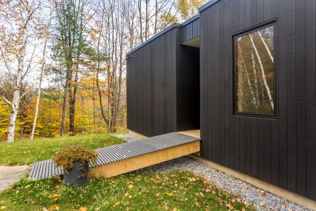 Exterior view of a contemporary dark-clad building set within a wooded autumn landscape. The angular single-level form features black vertical cladding, a recessed entry, a large dark-framed window, and a raised corrugated metal walkway leading toward the entrance. Gravel borders and grass surround the structure.