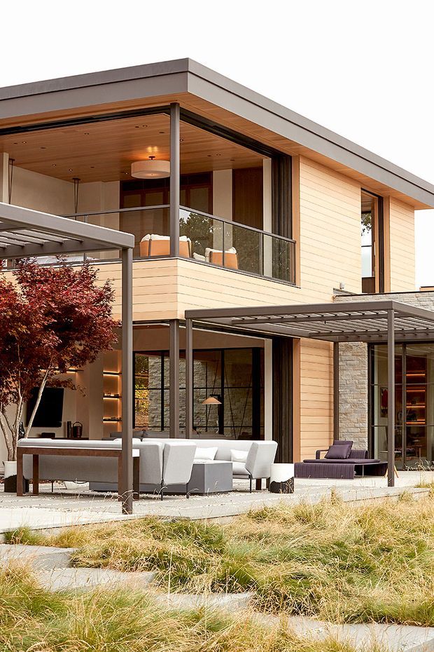 A contemporary multi-level residence viewed from the landscaped exterior courtyard. The home features a flat roof, pale horizontal cladding, extensive floor-to-ceiling glazing, dark slender frames, timber-lined soffits, stone wall sections, and covered outdoor living areas furnished with dining and lounge seating. A glazed balcony projects over the ground-floor terrace, while ornamental trees and grasses soften the modern architecture.