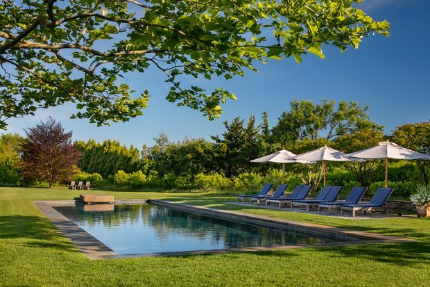 A serene landscaped outdoor swimming pool set within a spacious garden, with a long rectangular pool, stone or concrete coping, rows of blue sun loungers, and large white umbrellas. Mature trees, manicured lawn, and dense planting surround the pool under a clear blue sky.