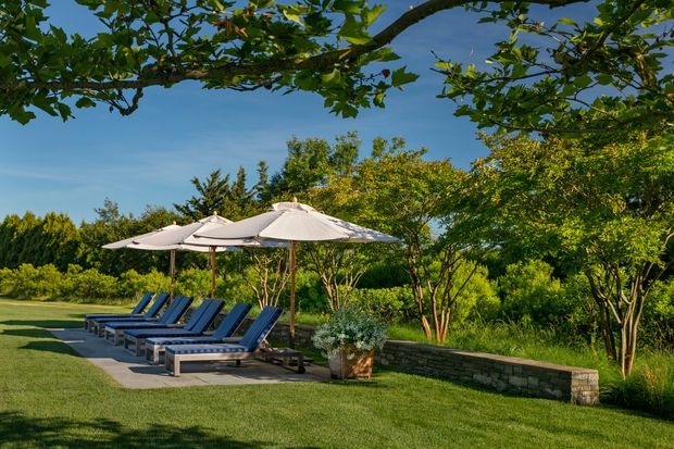 A landscaped outdoor lounging area with several blue-cushioned sun loungers arranged on a paved terrace beneath large white umbrellas. A low stone retaining wall, potted planting, manicured lawn, mature trees, and dense garden vegetation create a secluded resort-like setting.