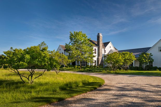 A broad exterior view of a large, traditional multi-gabled residence or estate surrounded by landscaped lawns and mature trees. The building features pale rendered or shingled walls, steep pitched gable roofs, dark roof coverings, a prominent stone chimney, and a long white-clad wing with a glazed roof section. A curved gravel driveway leads toward the house under clear daylight.