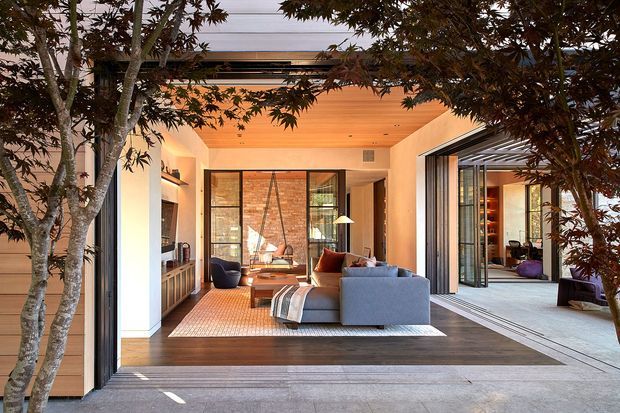 A warm, contemporary indoor-outdoor living room viewed from a covered terrace or courtyard. The room features a large grey sectional sofa, timber flooring, a pale textured rug, built-in timber cabinetry, expansive black-framed sliding glass doors, and a timber-lined ceiling. A glazed opening connects to an additional furnished space, while mature trees frame the foreground.