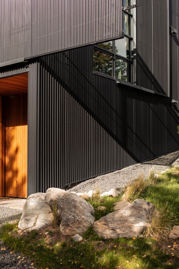 Close architectural exterior view of a dark, vertically ribbed metal-clad building with sharp intersecting volumes, black-framed windows, and a warm timber entry door. Strong sunlight creates dramatic shadows across the facade. A gravel edge, boulders, and grasses form a natural landscaped foreground.