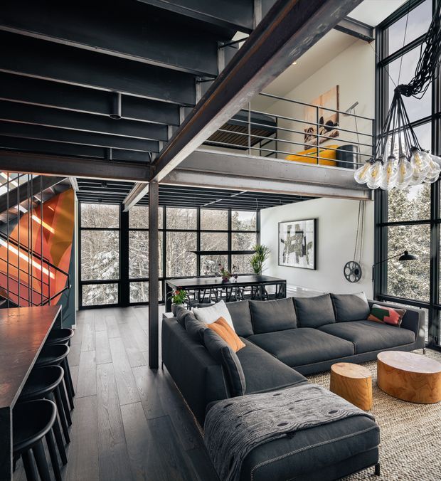 A dramatic double-height industrial-style living room with exposed steel beams, a mezzanine level, black-framed floor-to-ceiling glazing, and a large charcoal sectional sofa. The open-plan space includes a dining area, a bar or kitchen counter with stools, sculptural wood coffee tables, a textured rug, and a striking cluster pendant light. An orange-toned feature wall and artwork add warmth against the predominantly dark, neutral palette.